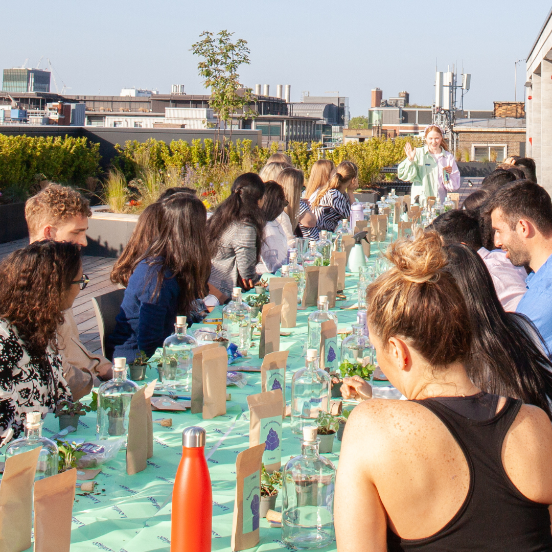 A group of people watching a woman demonstrate something on a table with various items.