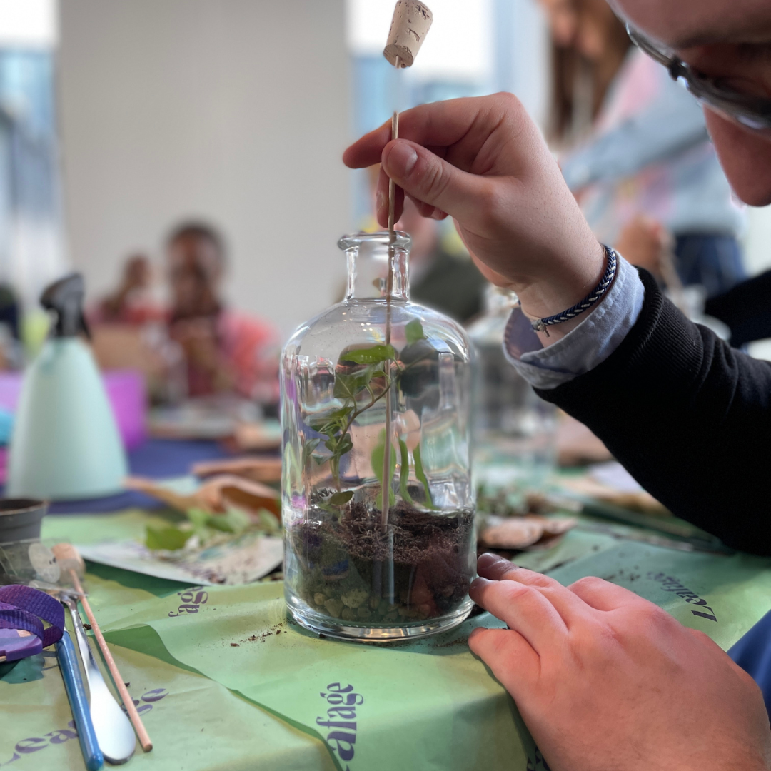 A group of people watching a woman demonstrate something on a table with various items.