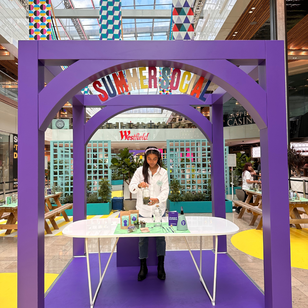 A group of people watching a woman demonstrate something on a table with various items.