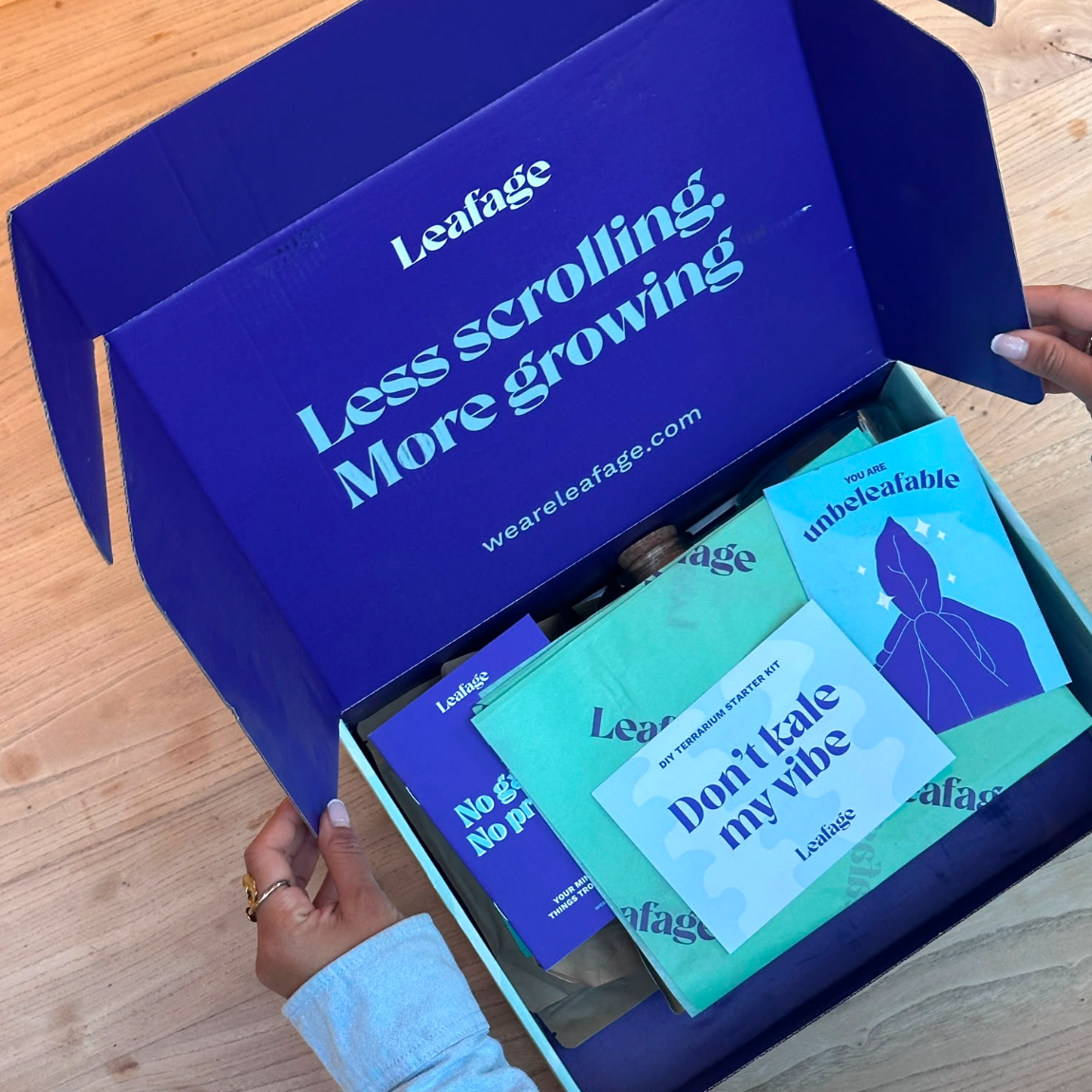 An open blue Leafage gift box on a light wood surface, showing several branded leaf-themed cards or packets inside. Hands are visible holding the box open, and the inside lid reads 'Leafage Less scrolling.